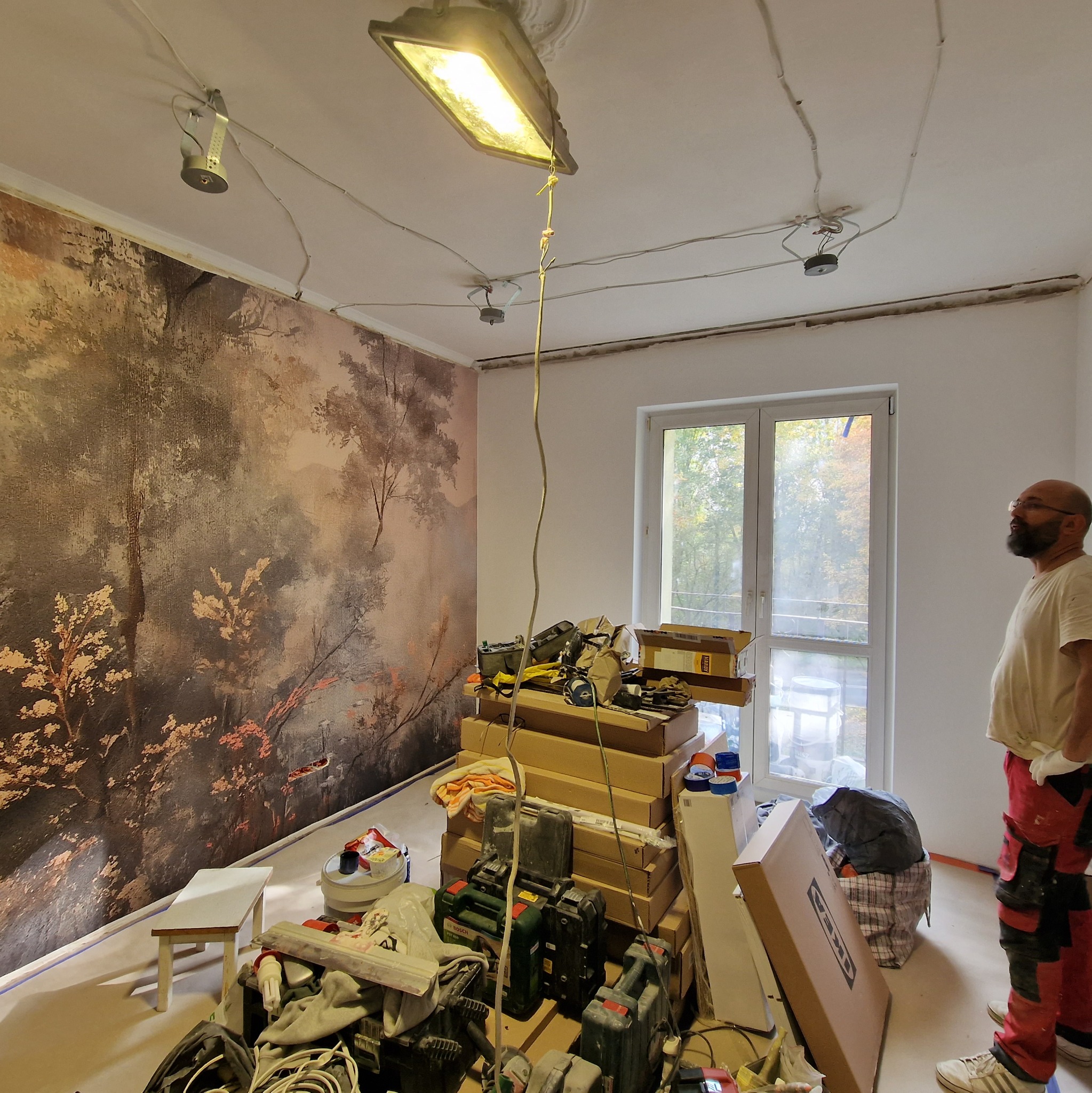 Pokój w trakcie remontu: ściana z fototapetą w leśny motyw, narzędzia i materiały budowlane zgromadzone na środku, widoczna instalacja oświetleniowa na suficie.