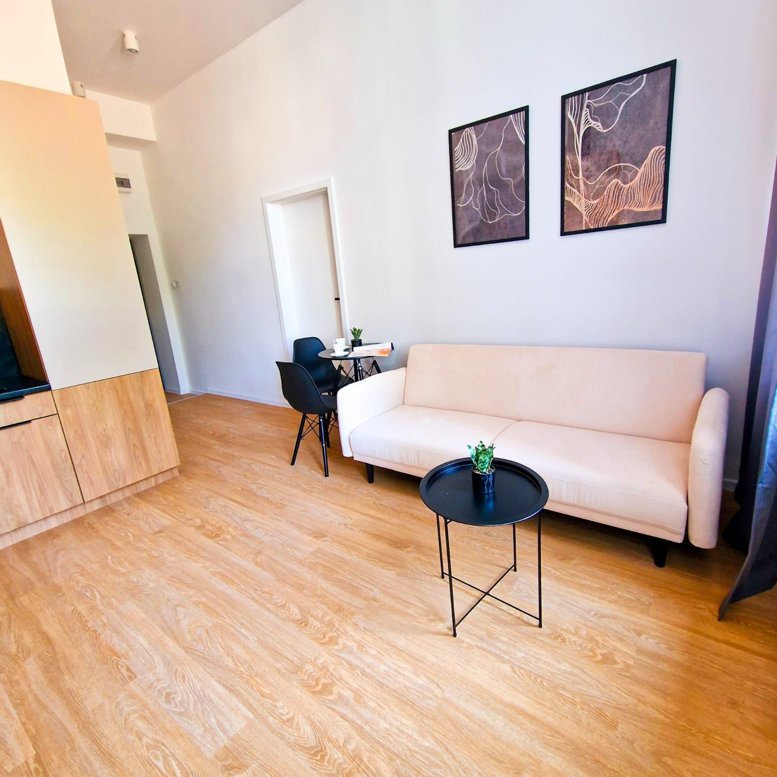 Jasne wnętrze salonu z drewnianą podłogą, beżową sofą, czarnym stolikiem i dekoracjami ściennymi. Widoczne meble i elementy wykończenia.