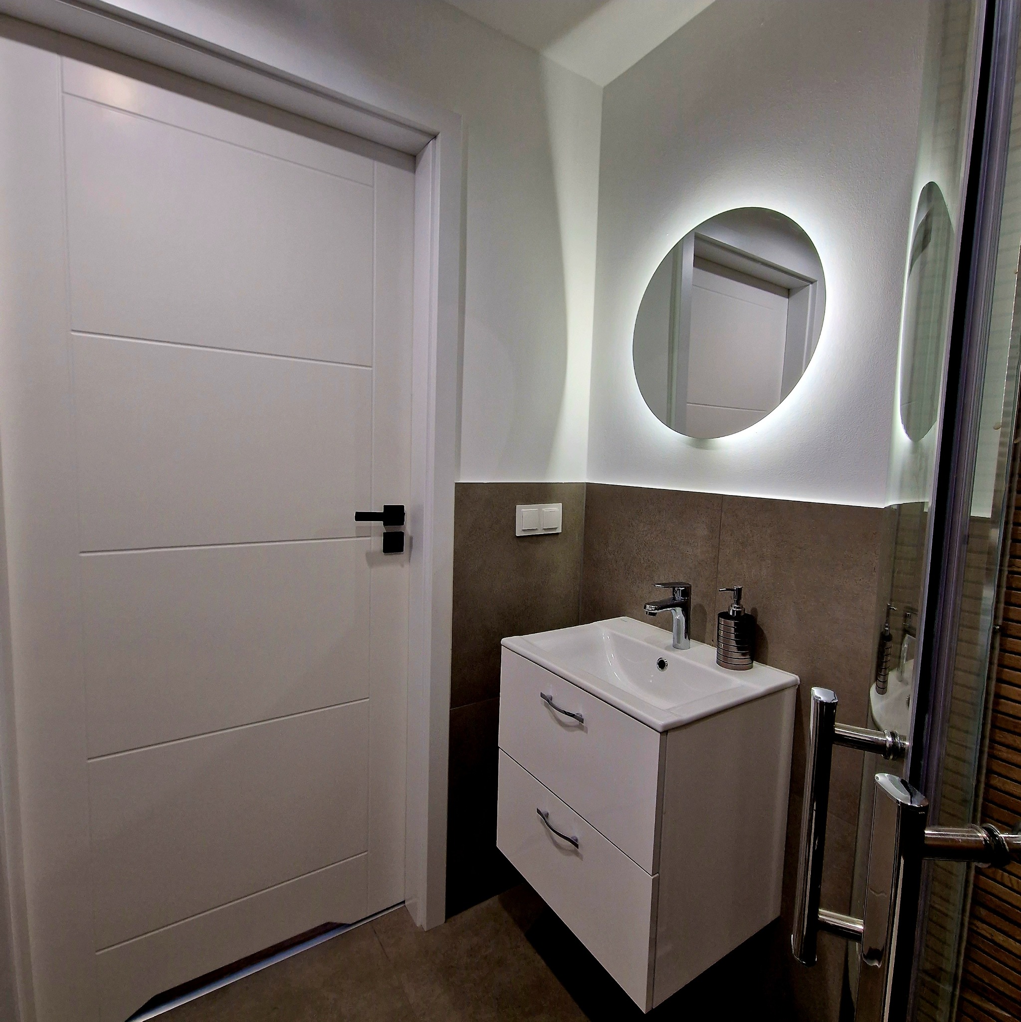 Nowoczesna łazienka z białą szafką pod umywalką, okrągłym lustrem z podświetleniem LED i częściowo widoczną kabiną prysznicową.