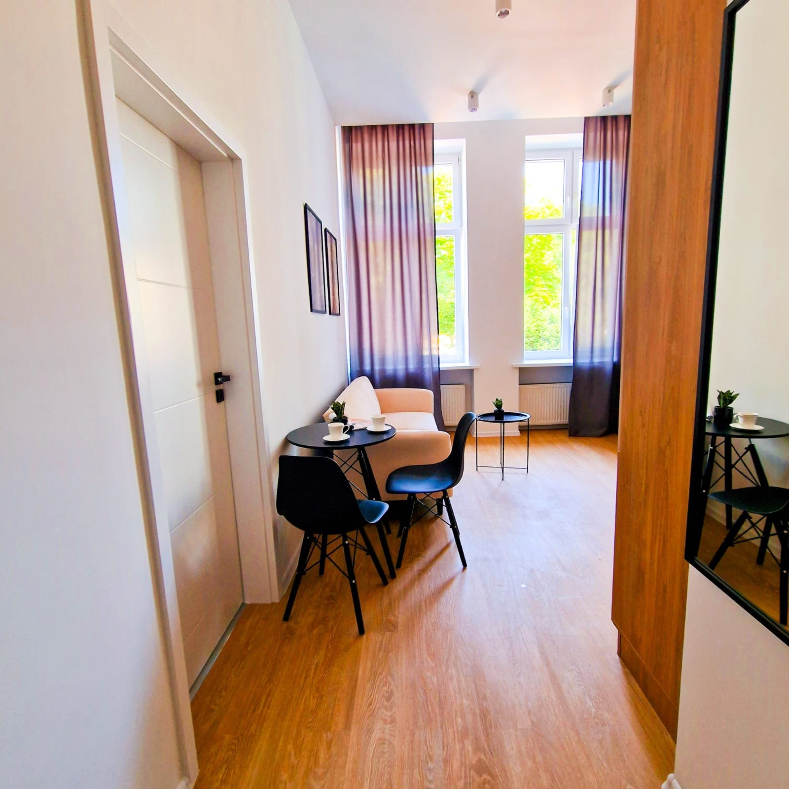 Jasne wnętrze pokoju z drewnianą podłogą, czarnymi krzesłami przy okrągłym stoliku i beżową sofą w tle, oświetlone naturalnym światłem z okien z fioletowymi zasłonami.