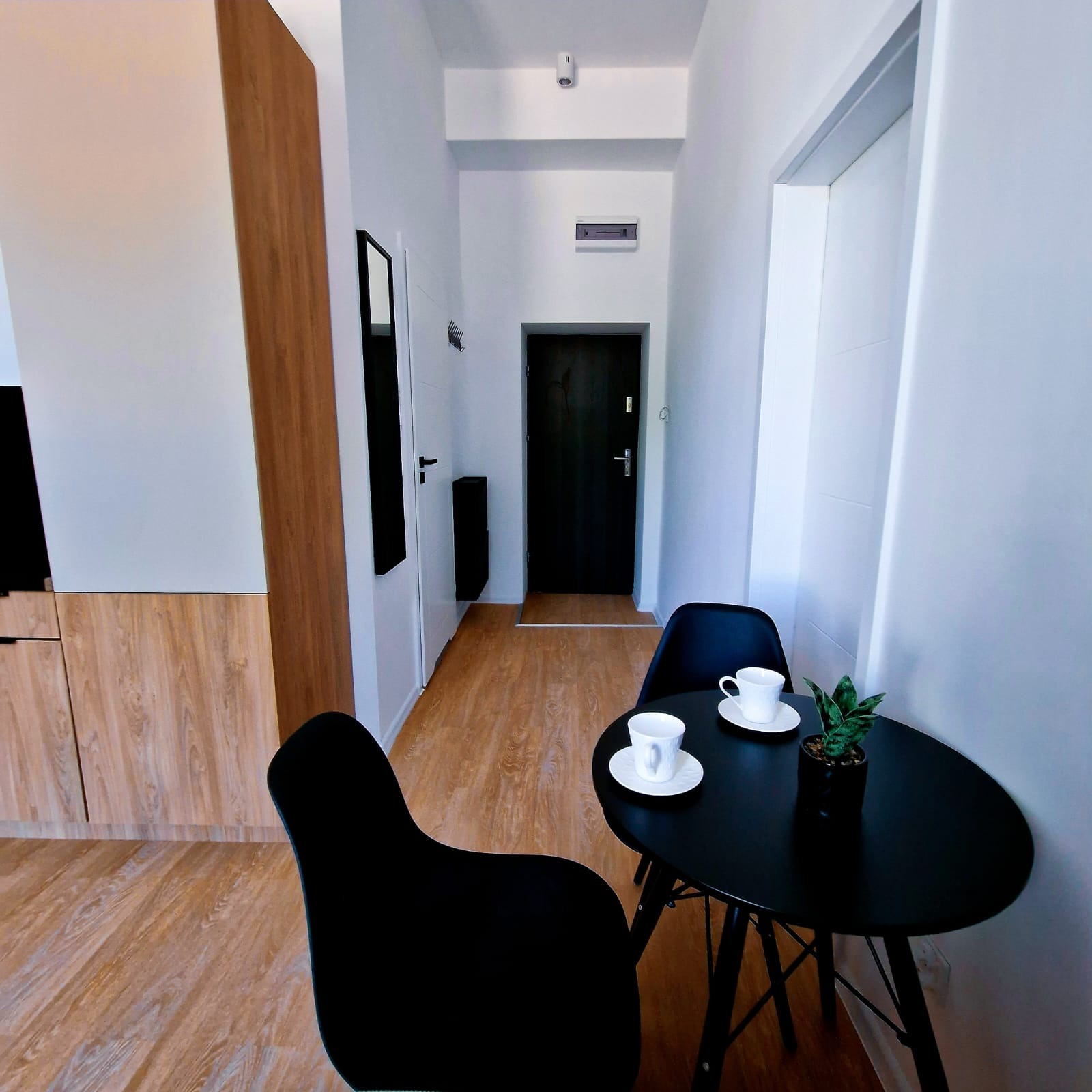 Nowoczesne wnętrze mieszkania: drewniana podłoga, białe ściany, czarne krzesła przy okrągłym stole z filiżankami i rośliną. Minimalistyczny design.