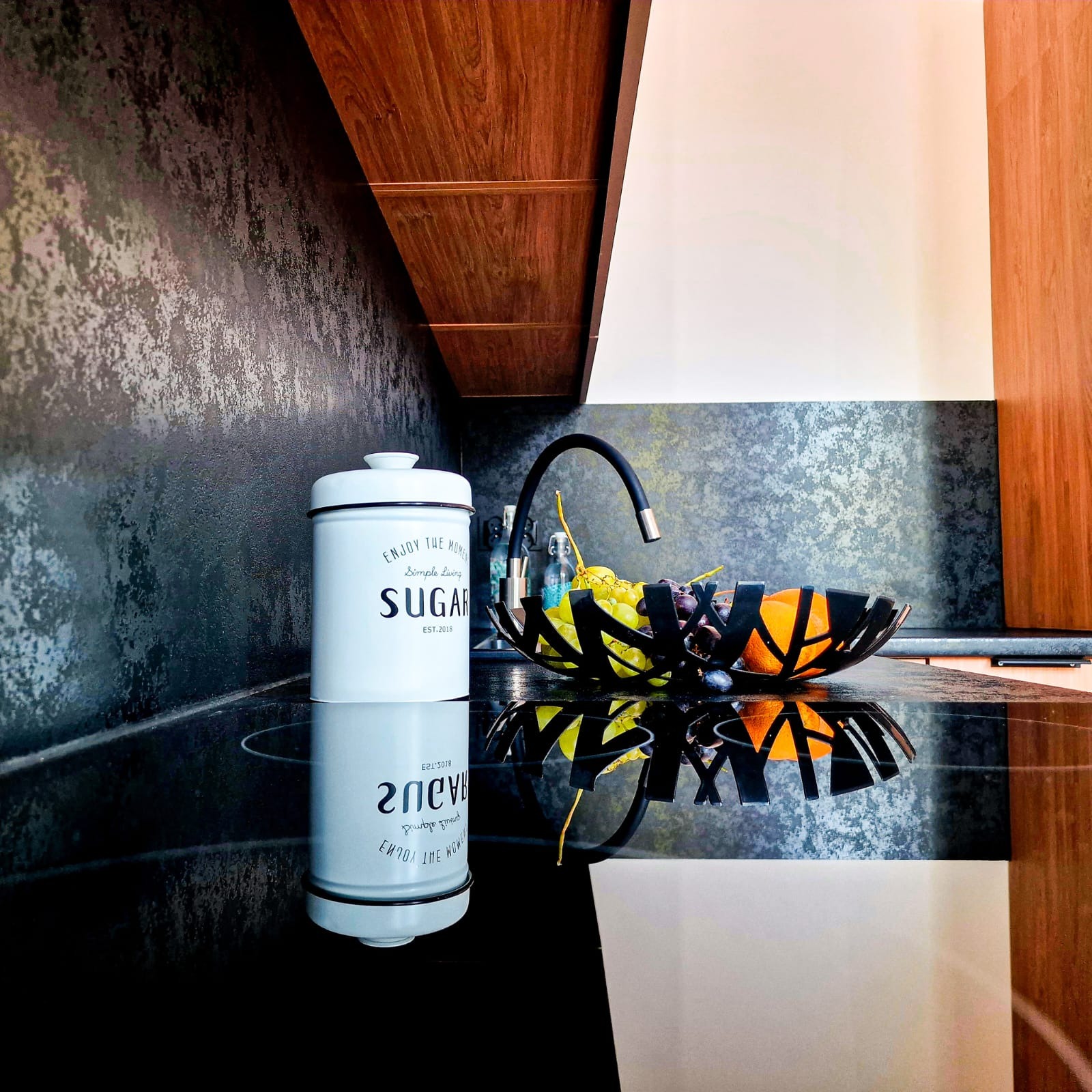 Nowoczesna kuchnia z blatem z czarnego kamienia, na którym stoi pojemnik na cukier i misa z owocami. W tle ściana z efektem betonu i drewniane szafki.