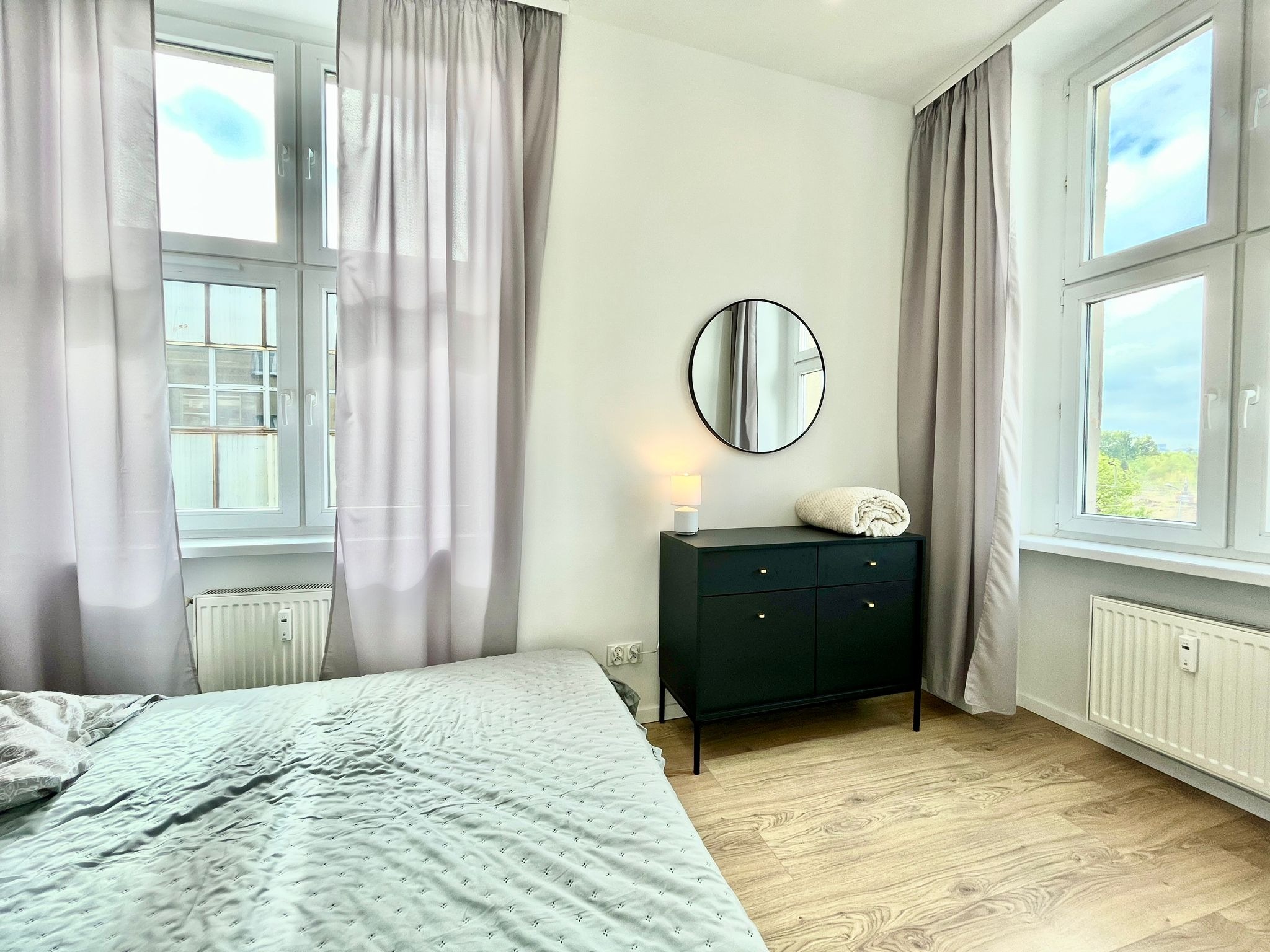 Jasne wnętrze sypialni z łóżkiem, komodą, okrągłym lustrem i oknami z zasłonami. Parkiet na podłodze. Minimalistyczny design.