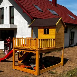 Łukasz Górka Usługi Stolarskie ŁG - Żółty drewniany domek dla dzieci ze zjeżdżalnią i piaskownicą na tle białego domu z czerwoną dachówką. Widoczni ludzie i zabawki.