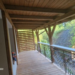 Łukasz Górka Usługi Stolarskie ŁG - Zadaszony taras z drewnianą podłogą i metalową balustradą o geometrycznym wzorze. Widok na zieleń i dach budynku w tle. Konstrukcja z belek.