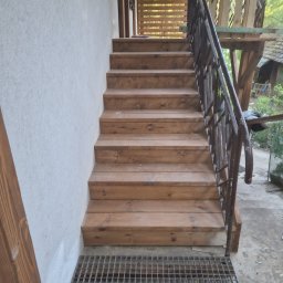Łukasz Górka Usługi Stolarskie ŁG - Drewniane schody zewnętrzne z metalową kratą u dołu i kutą balustradą, prowadzące do drewnianej konstrukcji. Widok z dołu, naturalne oświetlenie.