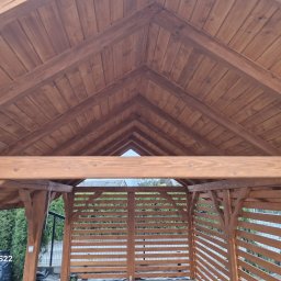 Łukasz Górka Usługi Stolarskie ŁG - Drewniana konstrukcja altany z widocznym dachem z desek, pomalowana na brązowo. Widok z wnętrza, z zewnątrz zieleń. Charakterystyczny wzór dachu.