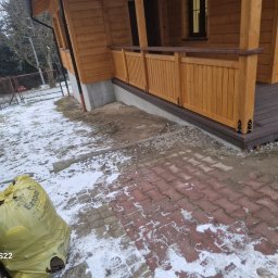 Łukasz Górka Usługi Stolarskie ŁG - Drewniany taras z balustradą przy domu z bali, widok z zewnątrz. Na pierwszym planie kostka brukowa i worek z odpadami budowlanymi. Zima, resztki śniegu.