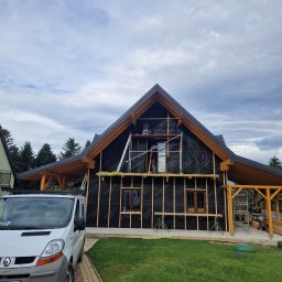 Łukasz Górka Usługi Stolarskie ŁG - Dom w trakcie ocieplania: widoczne rusztowania, izolacja czarną folią i drewniane listwy. Na pierwszym planie biały bus. Etap prac przed elewacją.