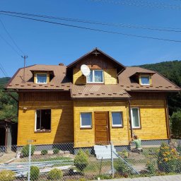 Łukasz Górka Usługi Stolarskie ŁG - Żółty, drewniany dom z brązowym dachem i satelitą. Osoba przy elewacji, prawdopodobnie maluje. Widoczne kwiaty i ogrodzenie.