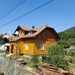 Łukasz Górka Usługi Stolarskie ŁG - Żółty dom z brązowym dachem w trakcie malowania elewacji przez pracownika na drabinie. W tle zielone wzgórza i błękitne niebo.