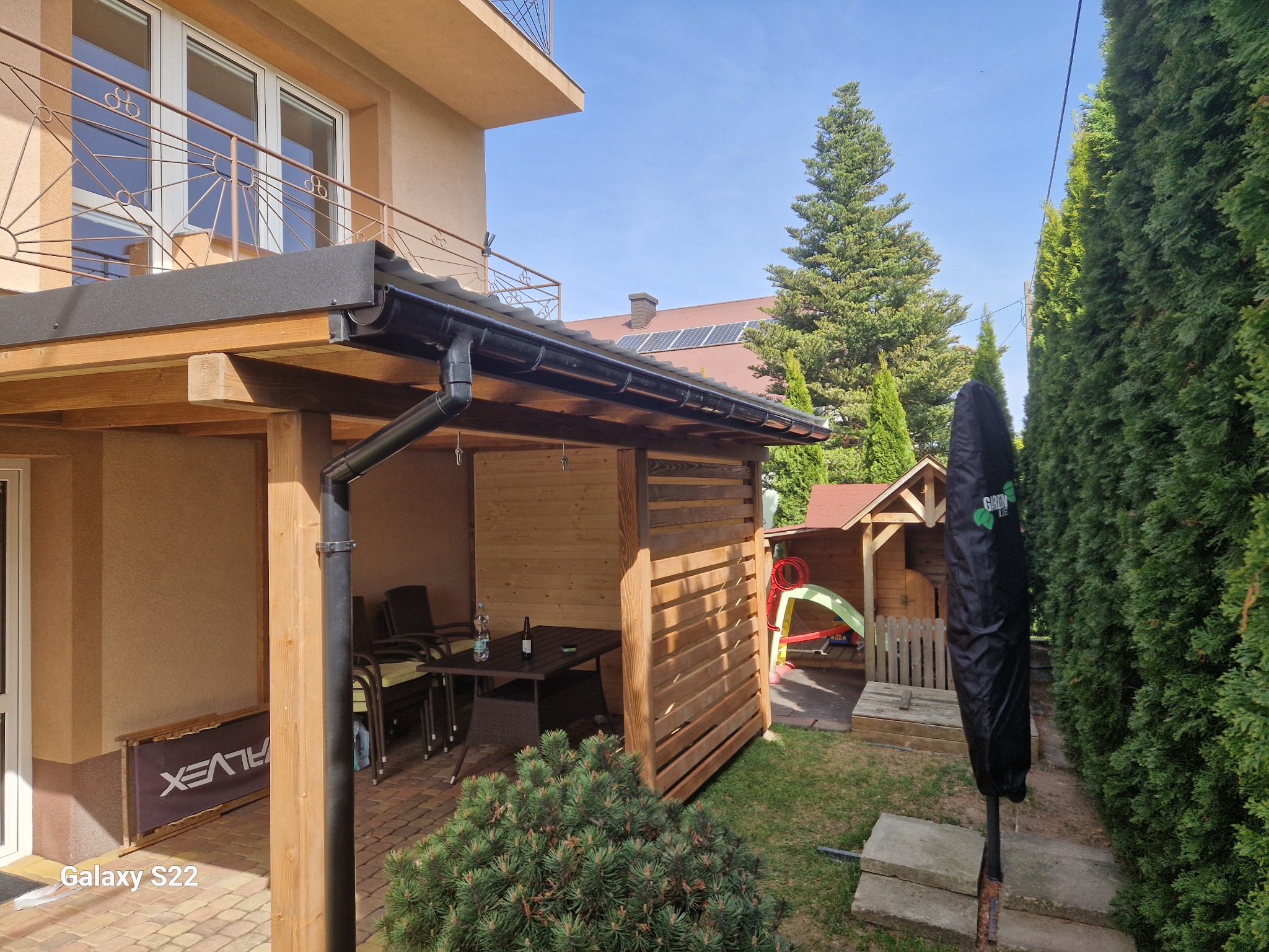 Zadaszony taras z drewnianą konstrukcją i czarną rynną, widok na ogród z domkiem dla dzieci i tujami. Na stole butelki, krzesła i stolik kawowy.