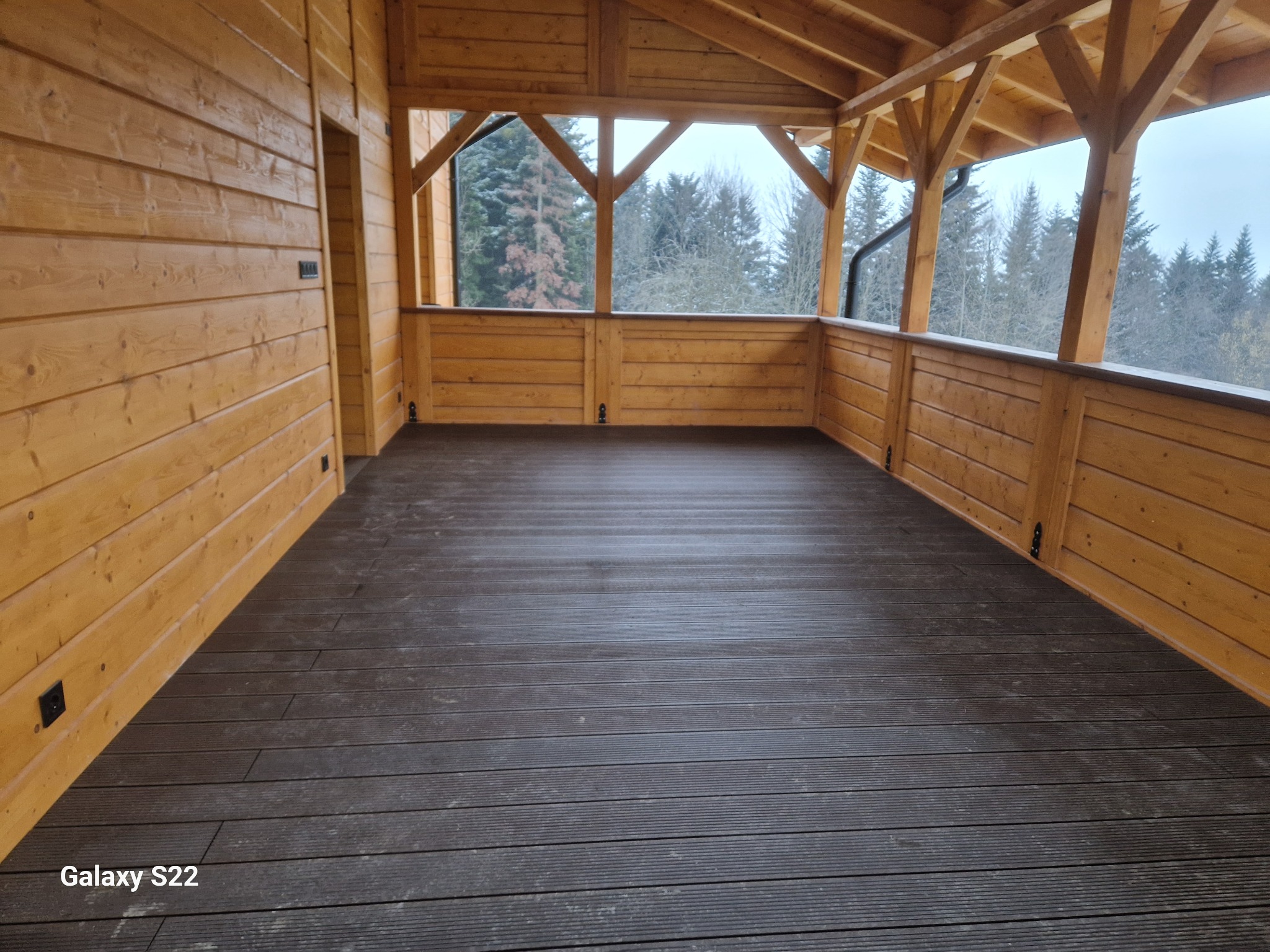 Zadaszony taras z drewnianą konstrukcją i kompozytową podłogą w odcieniach brązu, widok na zalesione wzgórza w tle. Widoczne gniazdka elektryczne.