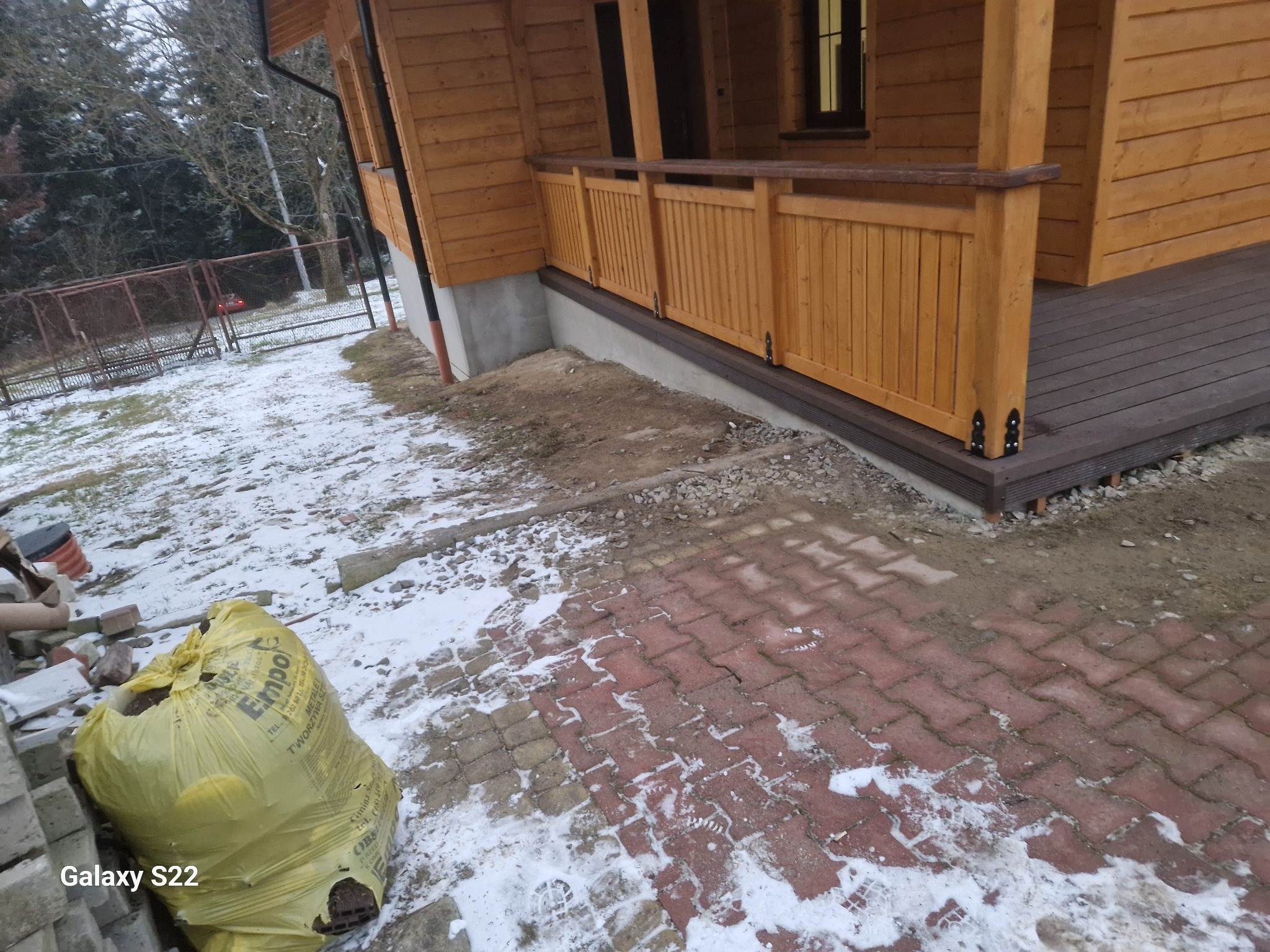Drewniany taras z balustradą przy domu z bali, widok z zewnątrz. Na pierwszym planie kostka brukowa i worek z odpadami budowlanymi. Zima, resztki śniegu.