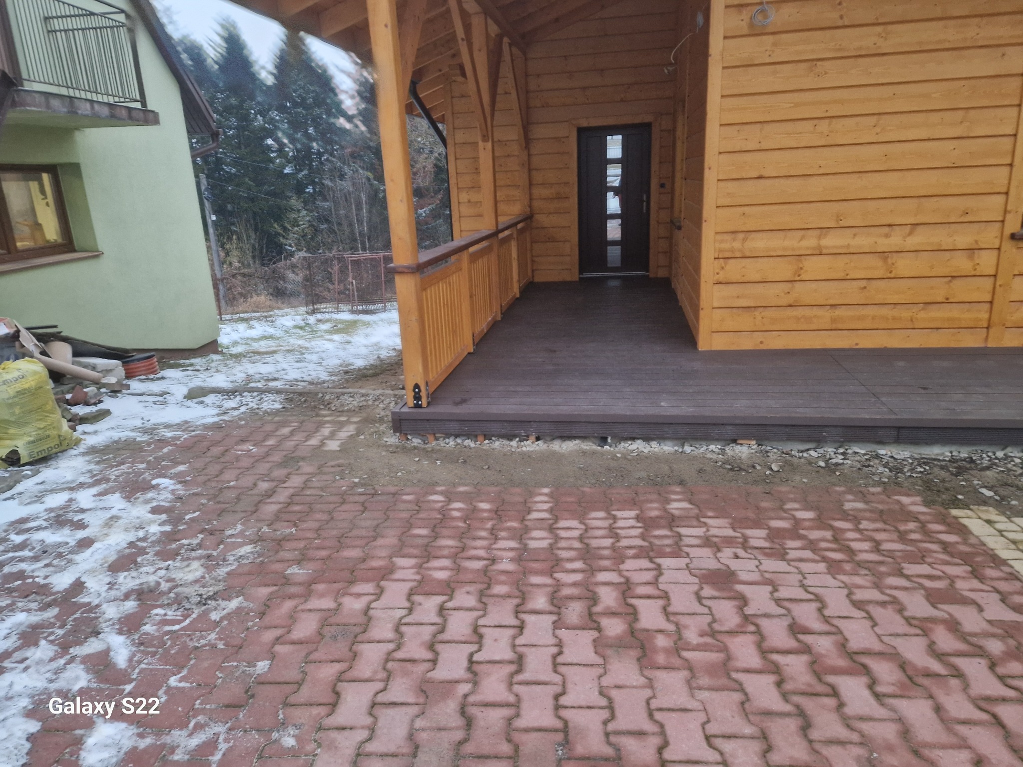 Drewniana altana z tarasem kompozytowym w zimowej scenerii. Widoczna kostka brukowa i fragment domu. Całość utrzymana w ciepłej kolorystyce drewna.