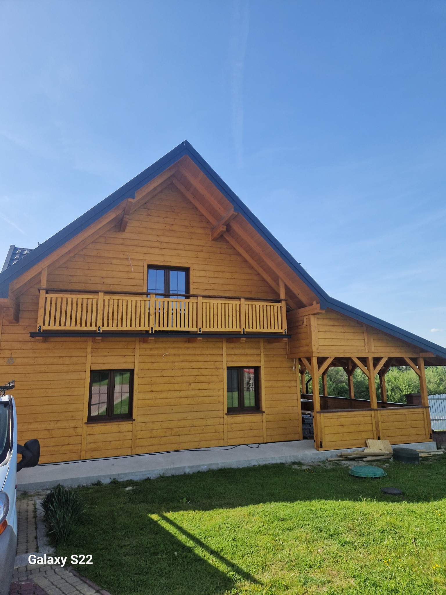 Żółty dom z drewna z balkonem i zadaszonym tarasem, ciemny dach, błękitne niebo. Widok z dołu, zieleń trawy w kadrze. Częściowo widoczny samochód.