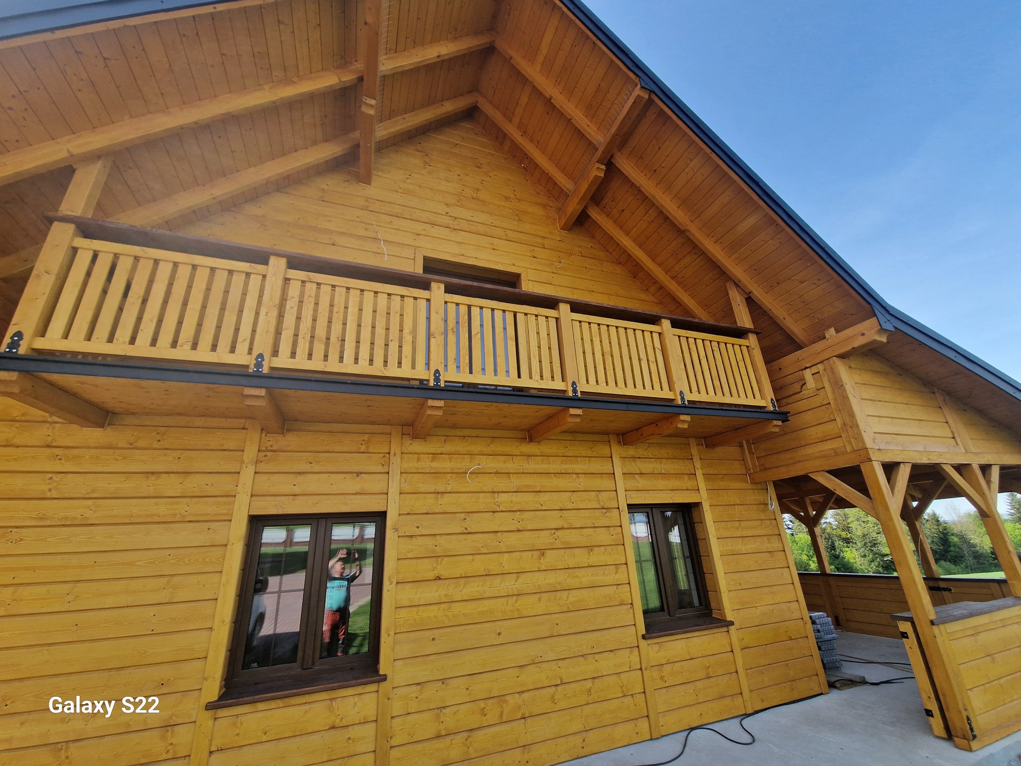 Drewniany dom z balkonem i zadaszonym tarasem, widok z dołu, błękitne niebo w tle. Widoczne detale konstrukcyjne i naturalna kolorystyka drewna.