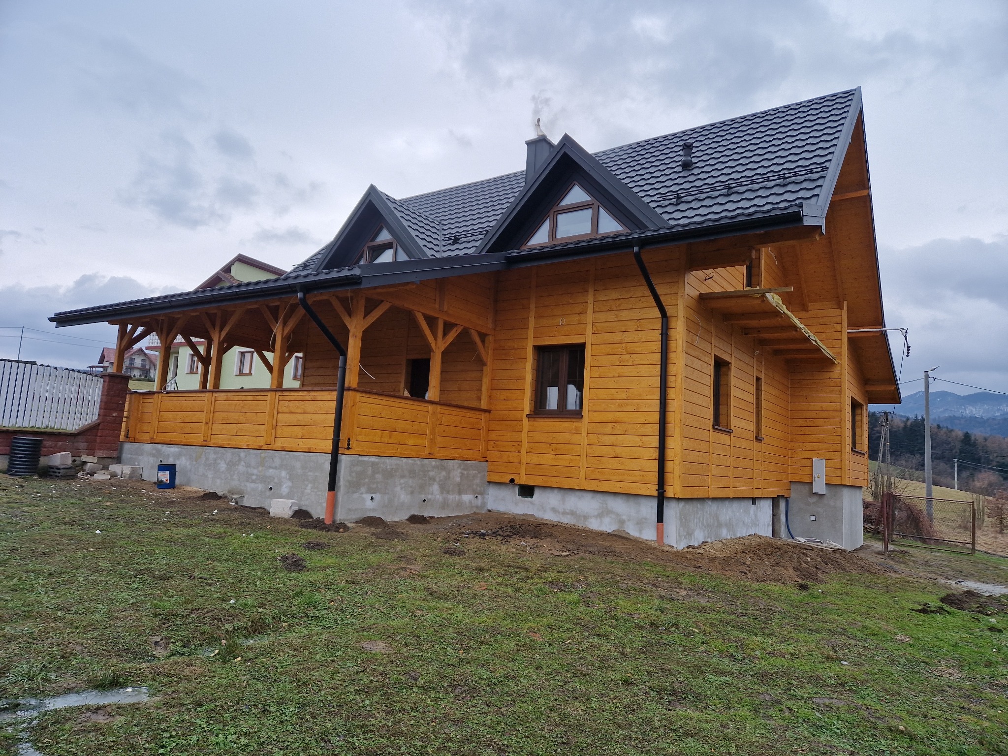 Drewniany dom z ciemną dachówką i rynnami, widok z zewnątrz. Elementy wykończeniowe w kolorze czarnym. Dom na tle pochmurnego nieba i gór.