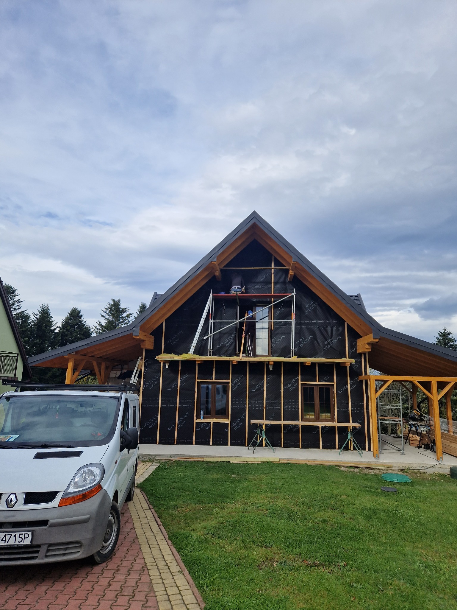 Dom w trakcie ocieplania: widoczne rusztowania, izolacja czarną folią i drewniane listwy. Na pierwszym planie biały bus. Etap prac przed elewacją.