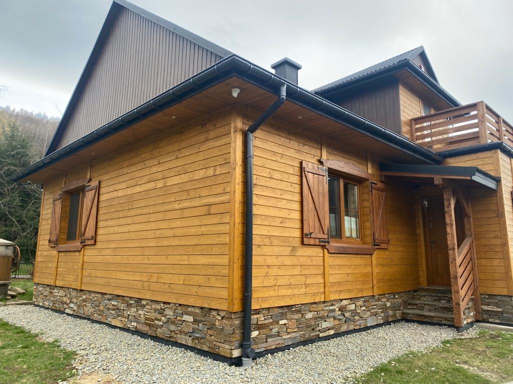 Drewniany dom z kamienną podmurówką, brązowymi okiennicami i czarnymi rynnami. Widok z boku, na tle zieleni i pochmurnego nieba. Detal: widoczna faktura drewna.