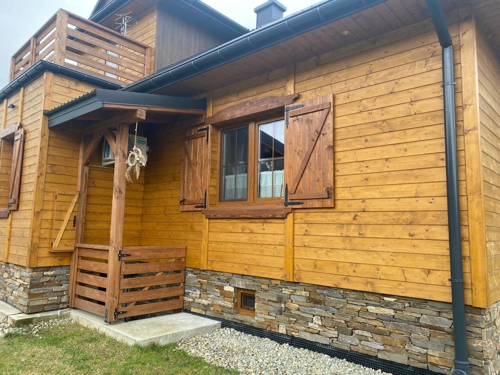 Drewniany dom z okiennicami i kamienną podmurówką, widok z zewnątrz. Czarna rynna pionowa na elewacji. Dekoracja nad wejściem.