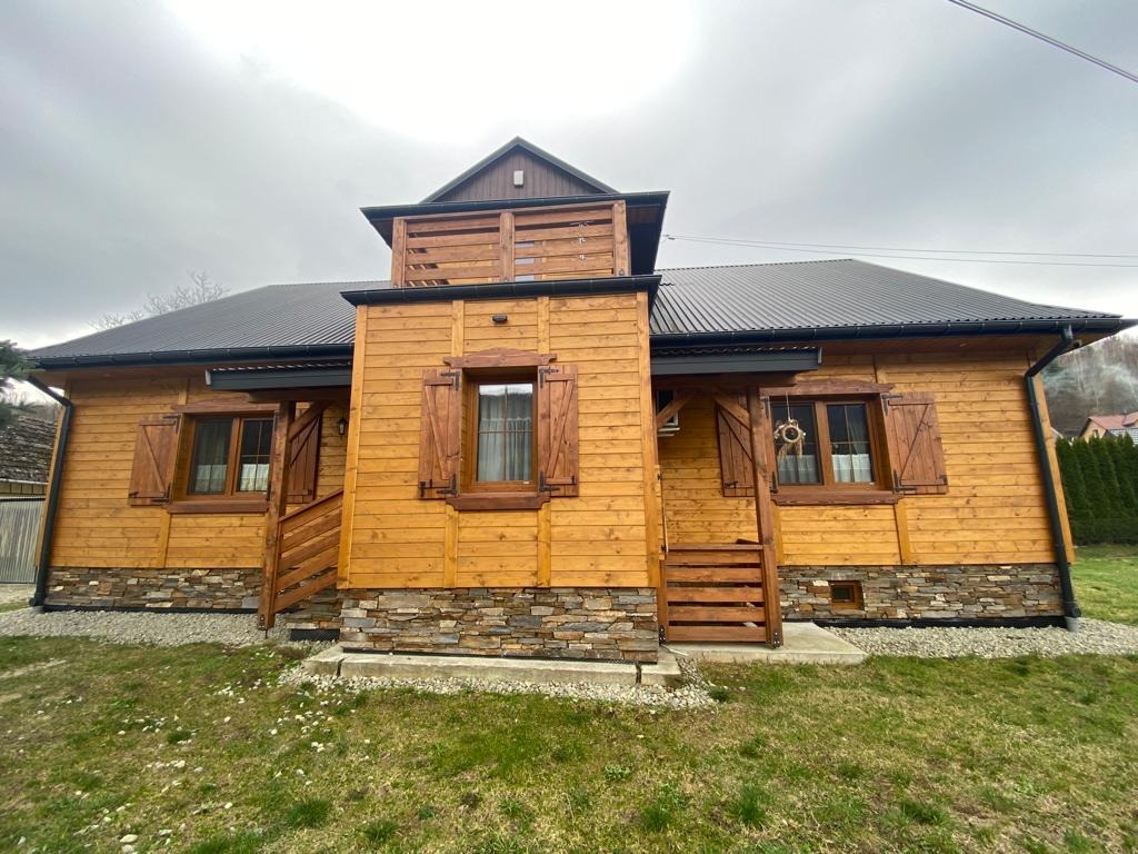 Drewniany dom z ciemnym dachem i okiennicami, wykończony kamieniem u podstawy. Widoczne rynny i schody wejściowe. Ujęcie z poziomu trawnika.