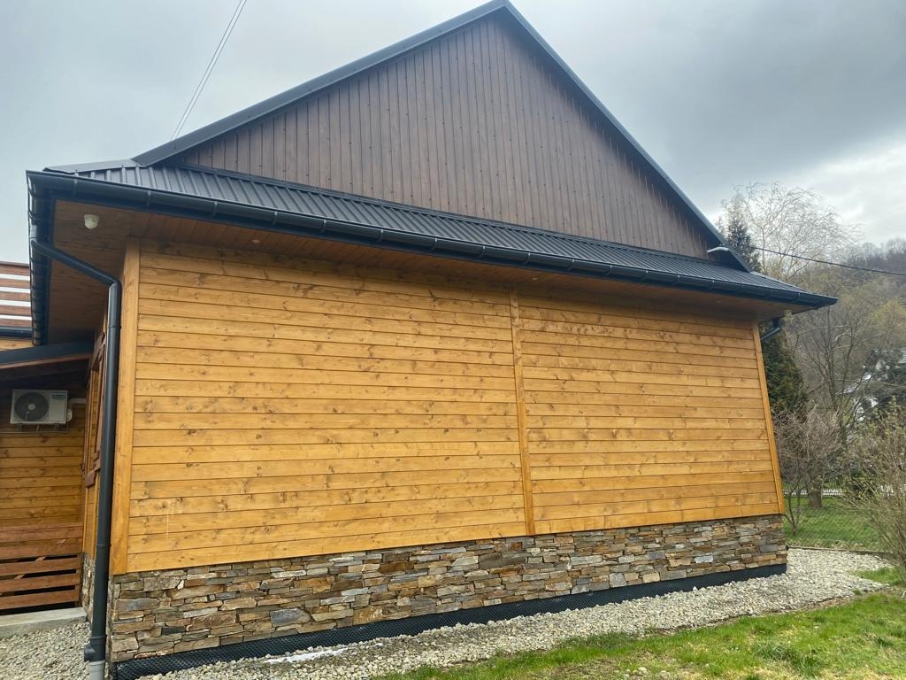 Drewniany dom z ciemnym dachem i rynnami, wykończony kamieniem u podstawy. Widok z boku, z fragmentem ogrodu i drzew w tle. Rynny w kolorze dachu.