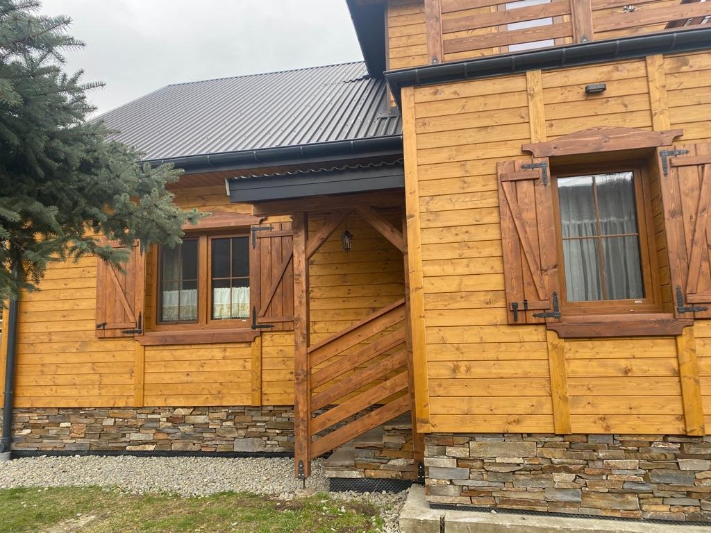 Drewniany dom z brązowymi okiennicami i kamienną podmurówką. Widoczny fragment dachu z metalowym pokryciem i rynny. Schody wejściowe z drewna. Całość w ciepłej tonacji.