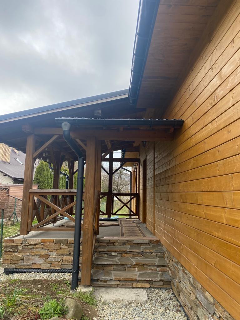 Drewniany dom z kamienną podmurówką i zadaszonym wejściem. Widoczne rynny i drewniana elewacja. Ujęcie z boku, podkreślające detale wykończenia.