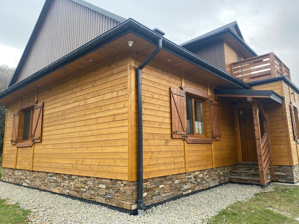 Drewniany dom z brązowymi rynnami, okiennicami i kamienną podmurówką. Widok z zewnątrz na elewację i fragment dachu. Uwagę przykuwa precyzyjny montaż rynien.