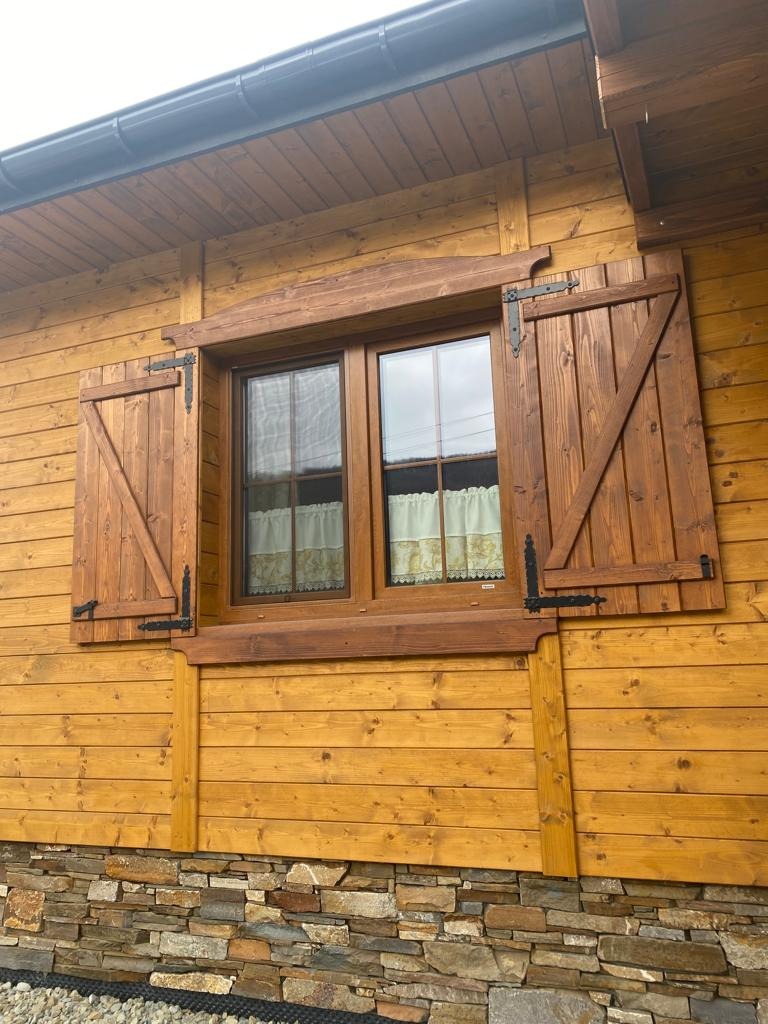 Drewniany dom z oknem i otwartymi okiennicami w rustykalnym stylu. Widoczne detale wykończenia, kamienna podmurówka i drewniana elewacja w ciepłym odcieniu.