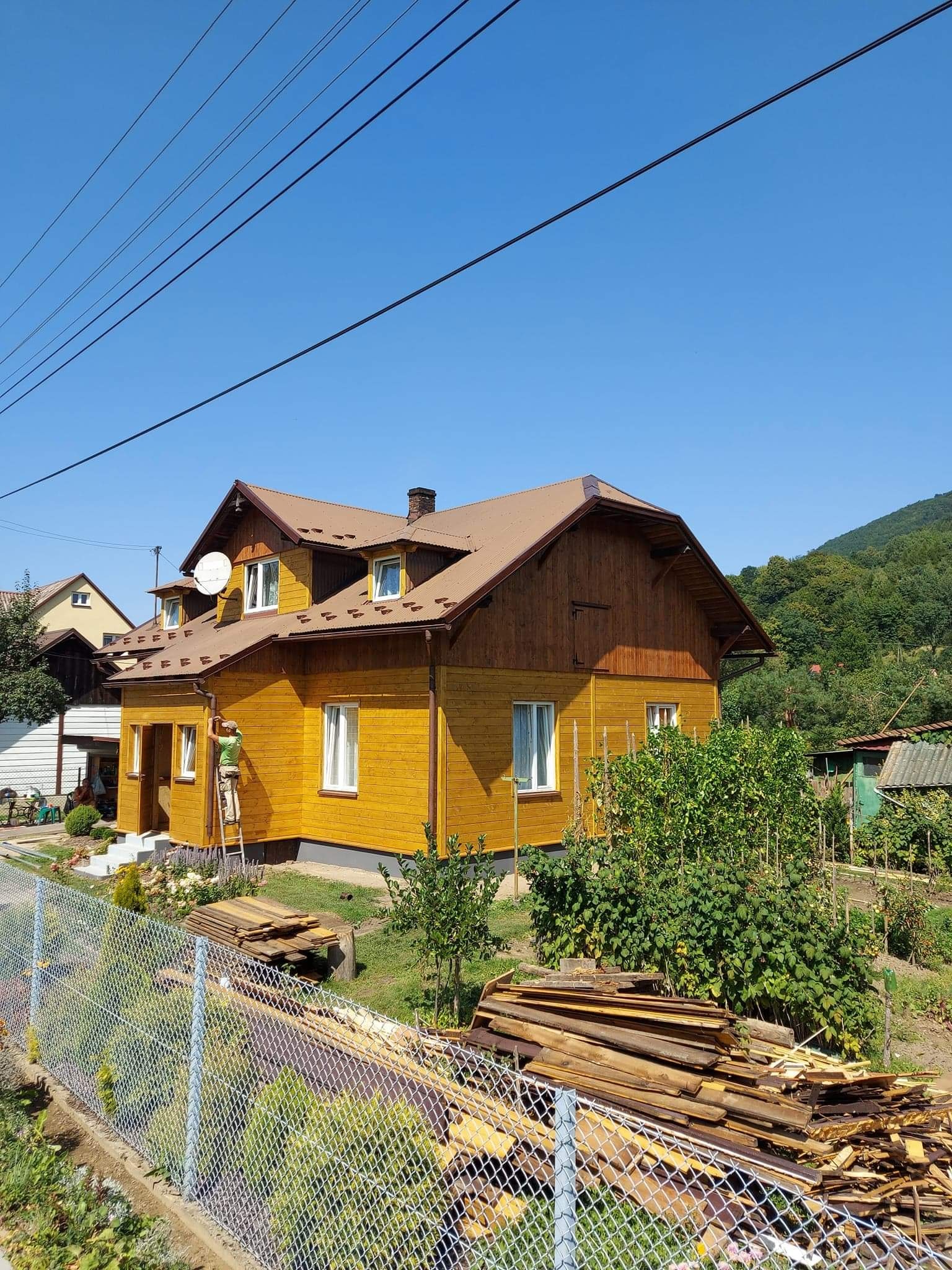 Żółty dom z brązowym dachem w trakcie malowania elewacji przez pracownika na drabinie. W tle zielone wzgórza i błękitne niebo.