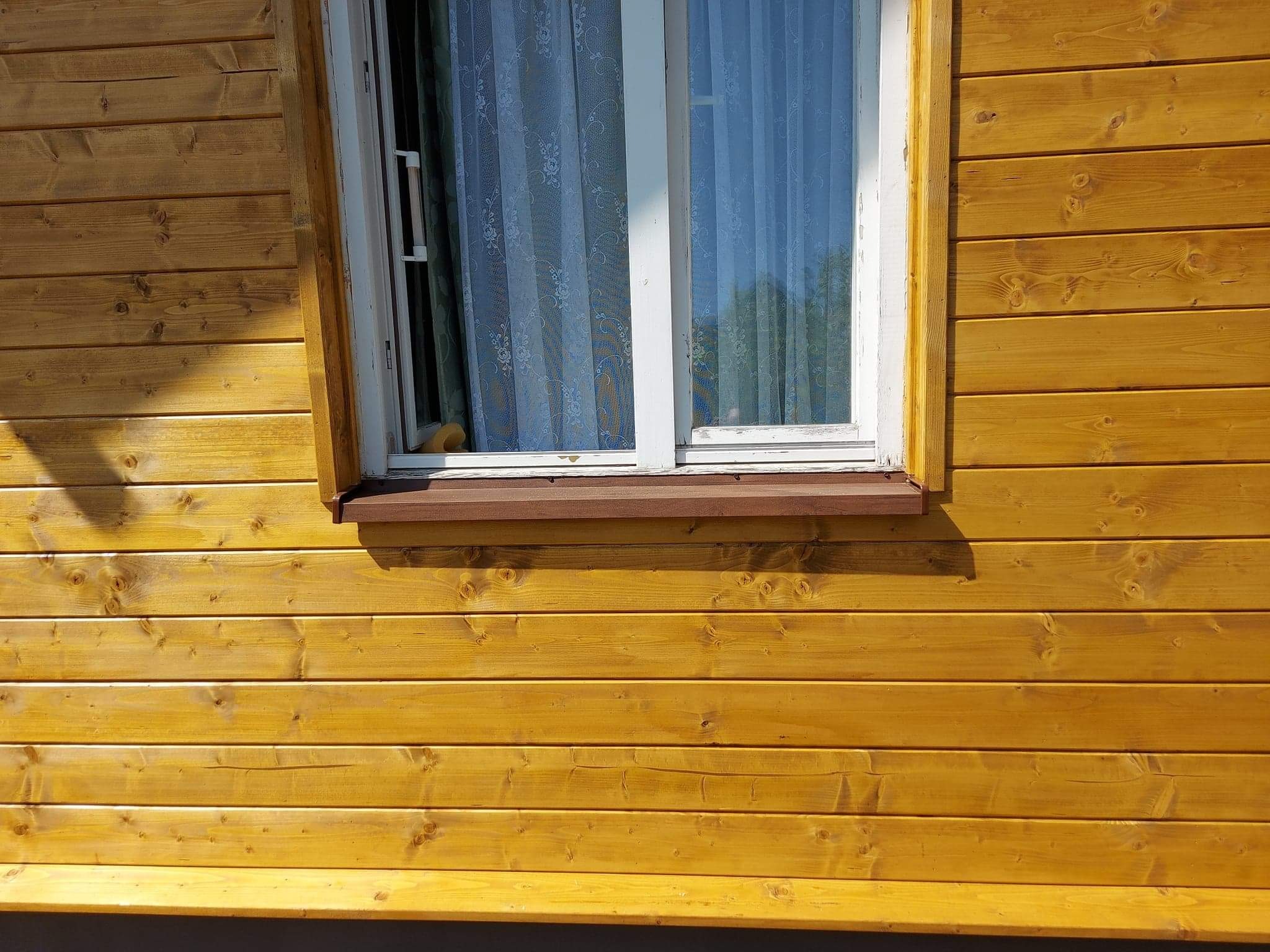 Żółta, drewniana elewacja domu z białym oknem i brązowym parapetem. Widoczne słoje drewna i delikatna firanka w oknie. Świeżo pomalowana powierzchnia.