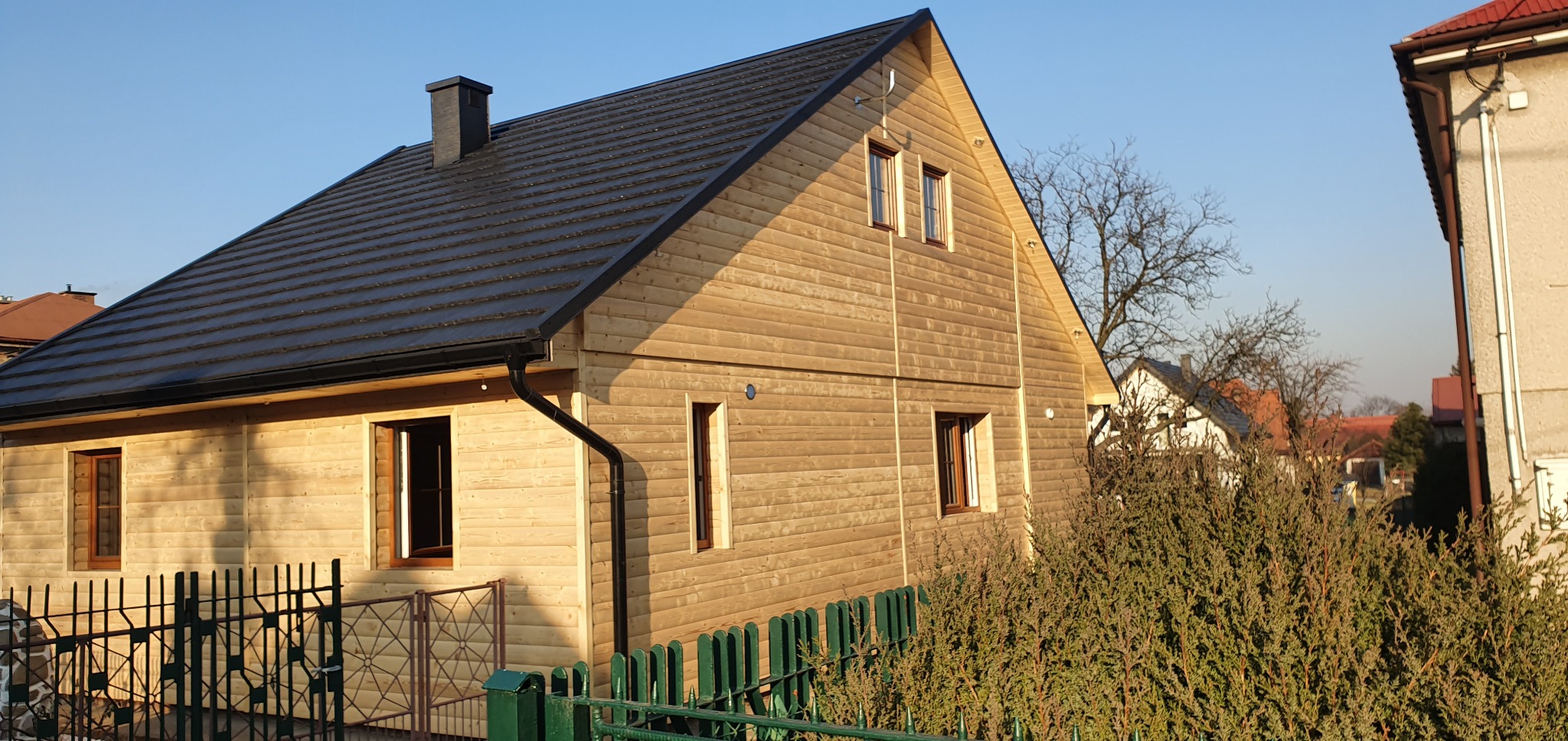 Nowy dom z drewnianą elewacją i ciemnym dachem, widok z zewnątrz, zielony płot w tle. Ujęcie w słoneczny dzień.