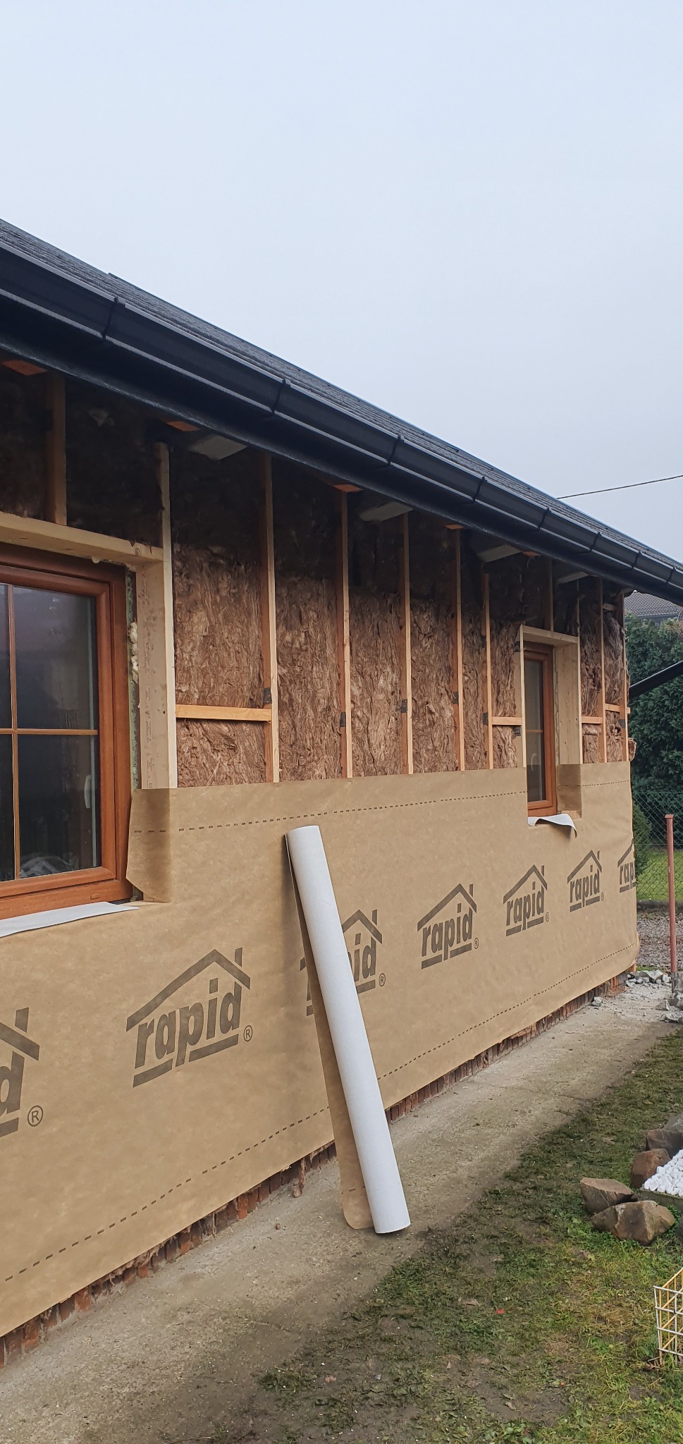 Ocieplenie budynku z widoczną izolacją i membraną wiatroizolacyjną z nadrukiem. Widoczne okna i fragment dachu z rynnami. Prace w toku, zwinięta rolka materiału oparta o ścianę.