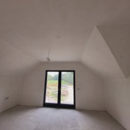 Mazbud - Poddasze po gładziowaniu ścian i wylewce posadzki, z widocznym oknem balkonowym i wystającymi przewodami elektrycznymi. Ściany i sufit w jasnym odcieniu bieli.