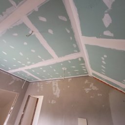 Mazbud - Skośny sufit z płyt kartonowo-gipsowych w trakcie szpachlowania, widoczne łączenia i ślady po wkrętach. W tle fragment ściany i ościeżnica drzwi.