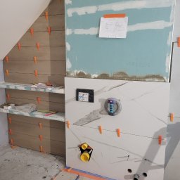 Mazbud - Układanie płytek imitujących drewno na skośnej ścianie w łazience, widoczne elementy instalacji i narzędzia glazurnicze, w tle płyta karton-gips.