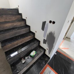 Mazbud - Schody z ciemnego marmuru z drewnianymi akcentami w trakcie wykańczania. Na ścianie widoczne próbki farb i przygotowane otwory pod gniazdka. Podłoga zabezpieczona kartonem.
