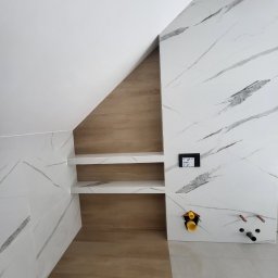 Mazbud - Wykończona wnęka z półkami w łazience, wyłożona płytkami imitującymi marmur i drewno. Widoczne przyłącza wodno-kanalizacyjne i panel sterowania.