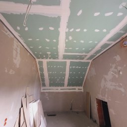 Mazbud - Sufit z płyt karton-gips, szpachlowane łączenia, widoczne kable, ściany w trakcie przygotowania do malowania. Prace wykończeniowe w pomieszczeniu ze skosami.
