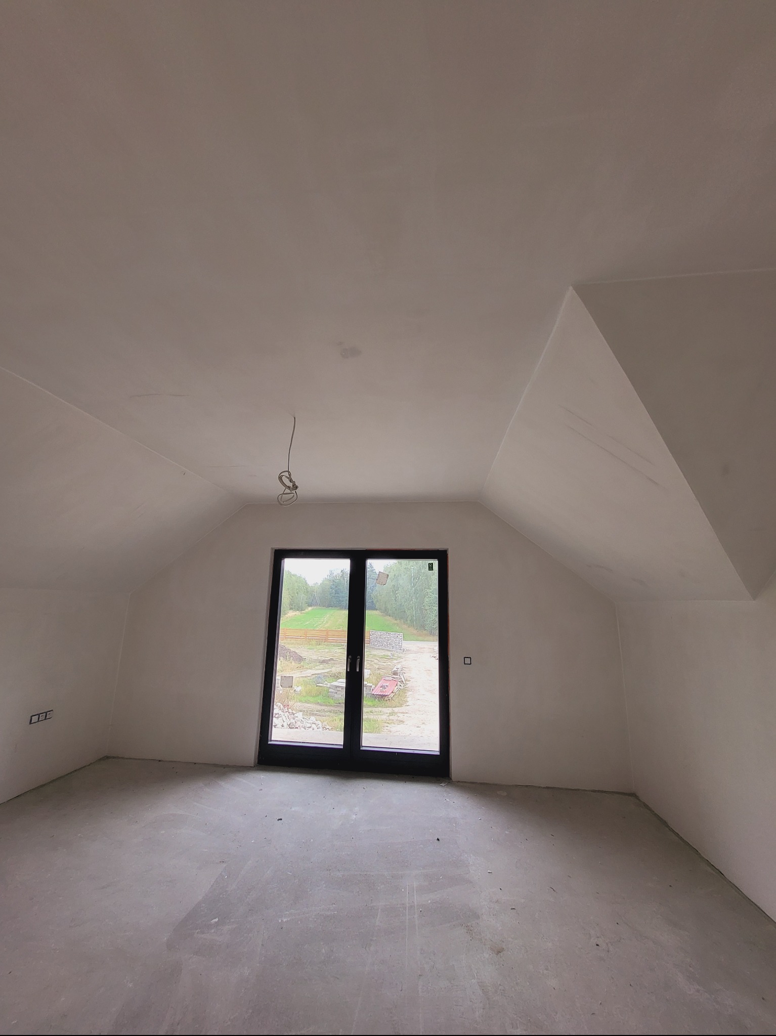 Poddasze po gładziowaniu ścian i wylewce posadzki, z widocznym oknem balkonowym i wystającymi przewodami elektrycznymi. Ściany i sufit w jasnym odcieniu bieli.