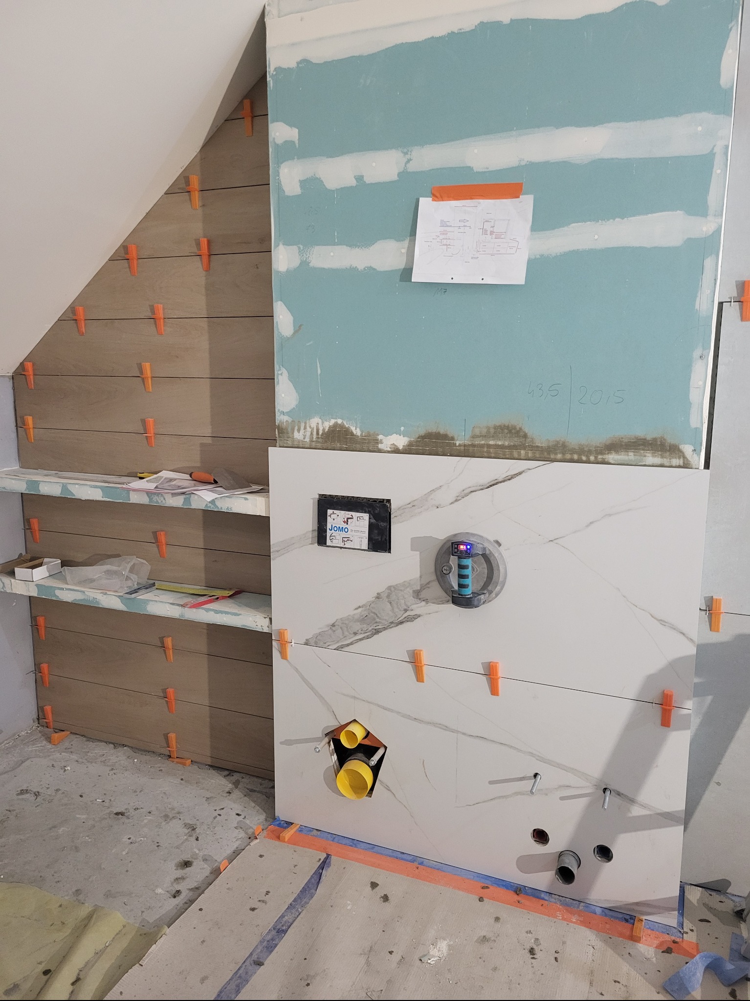 Układanie płytek imitujących drewno na skośnej ścianie w łazience, widoczne elementy instalacji i narzędzia glazurnicze, w tle płyta karton-gips.