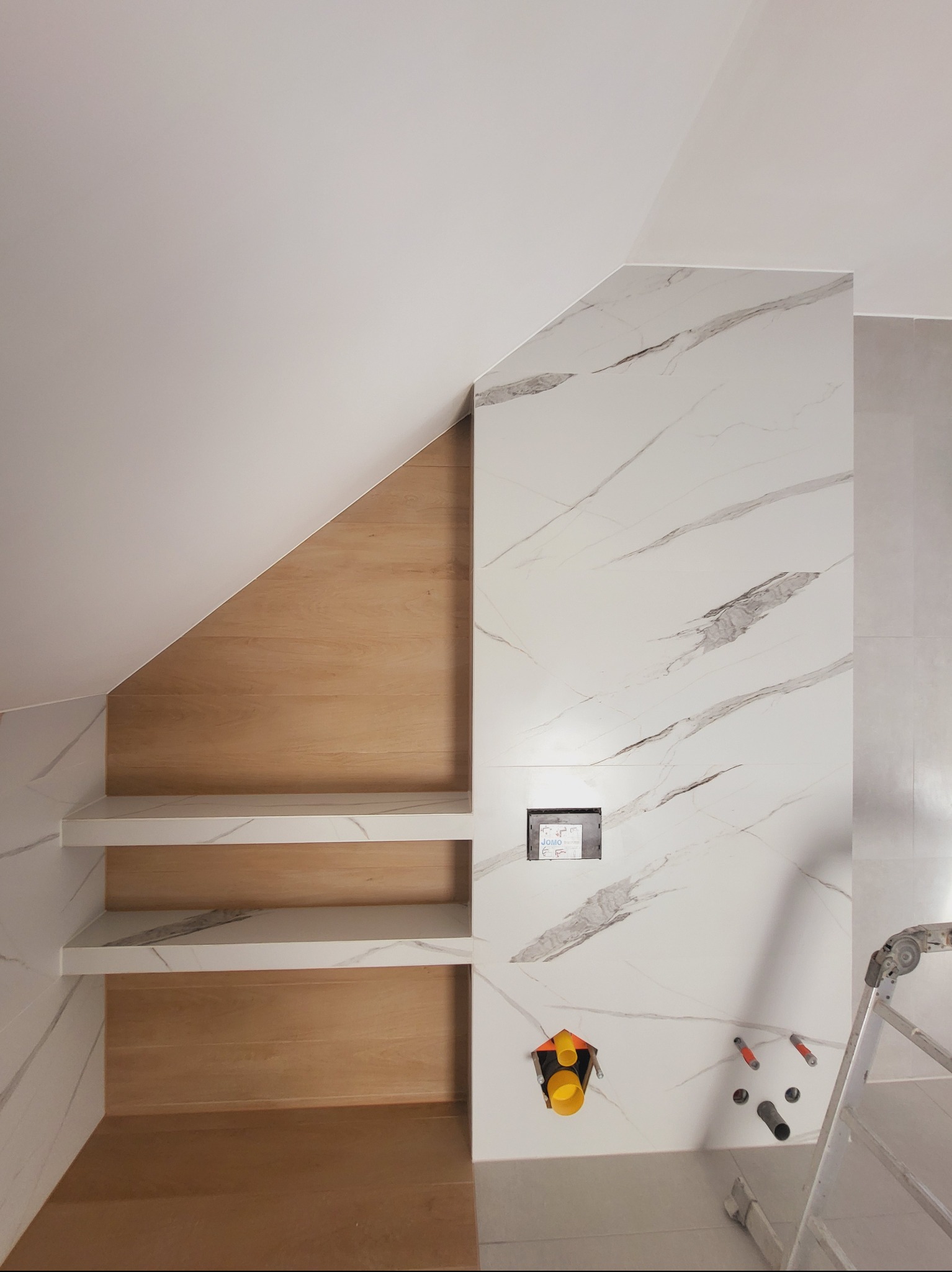 Wykończona wnęka z półkami w łazience, wyłożona płytkami imitującymi marmur i drewno. Widoczne przyłącza wodno-kanalizacyjne i drabina.