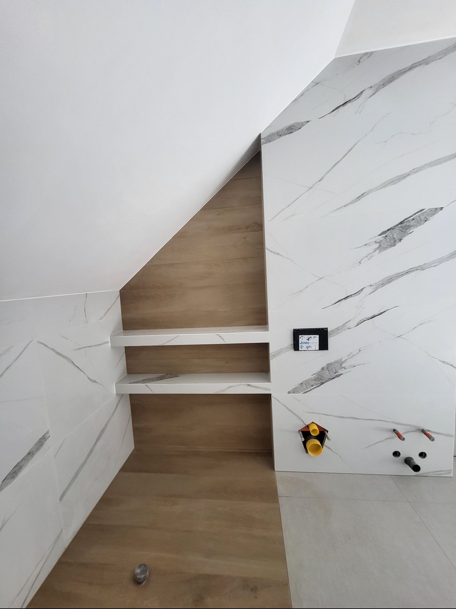 Wykończona wnęka z półkami w łazience, wyłożona płytkami imitującymi marmur i drewno. Widoczne przyłącza wodno-kanalizacyjne i panel sterowania.