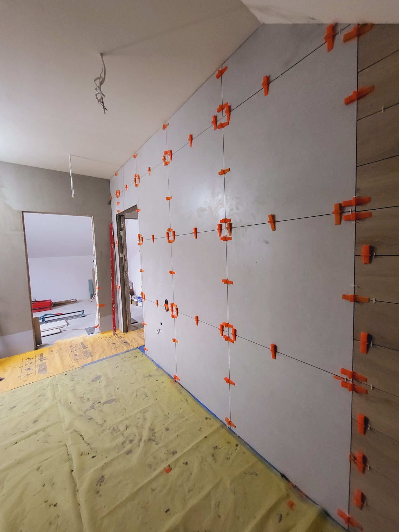Ściana w trakcie układania płytek z użyciem pomarańczowych klipsów poziomujących. Widoczne sznurki wyznaczające linie. Podłoga zabezpieczona folią ochronną.