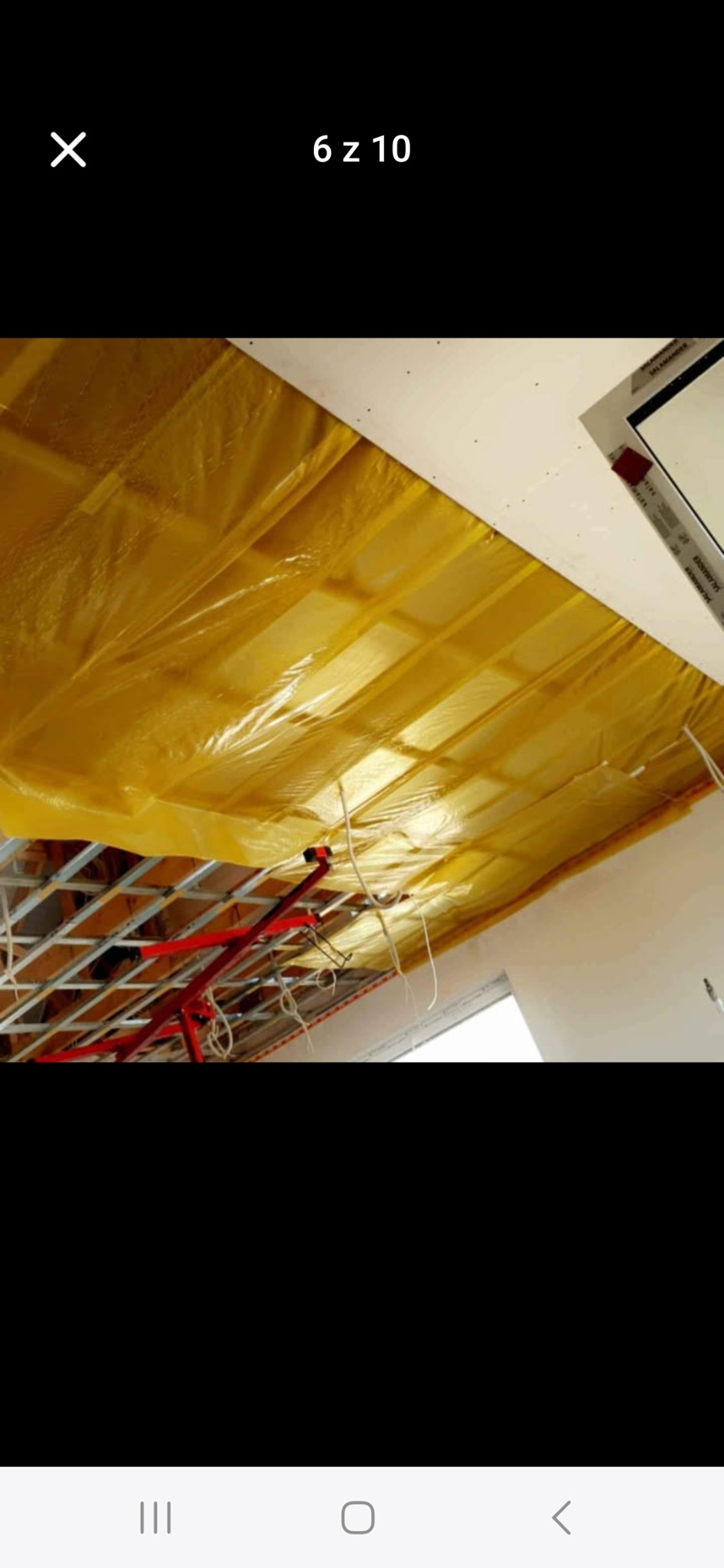 Żółta folia zabezpieczająca na metalowej konstrukcji sufitu podwieszanego, widoczne kable i fragment ściany. Etap prac przed montażem płyt.