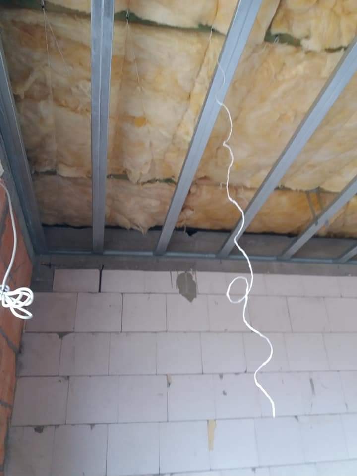 Montaż izolacji z wełny mineralnej w metalowym stelażu sufitu podwieszanego, widok z dołu na surową konstrukcję i fragment ściany z bloczków.