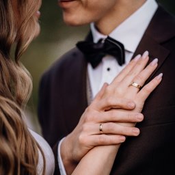 Paweł Liczbik - Zbliżenie dłoni pary młodej z obrączkami na tle garnituru i sukni. Delikatne światło podkreśla detale, tworząc intymną atmosferę.