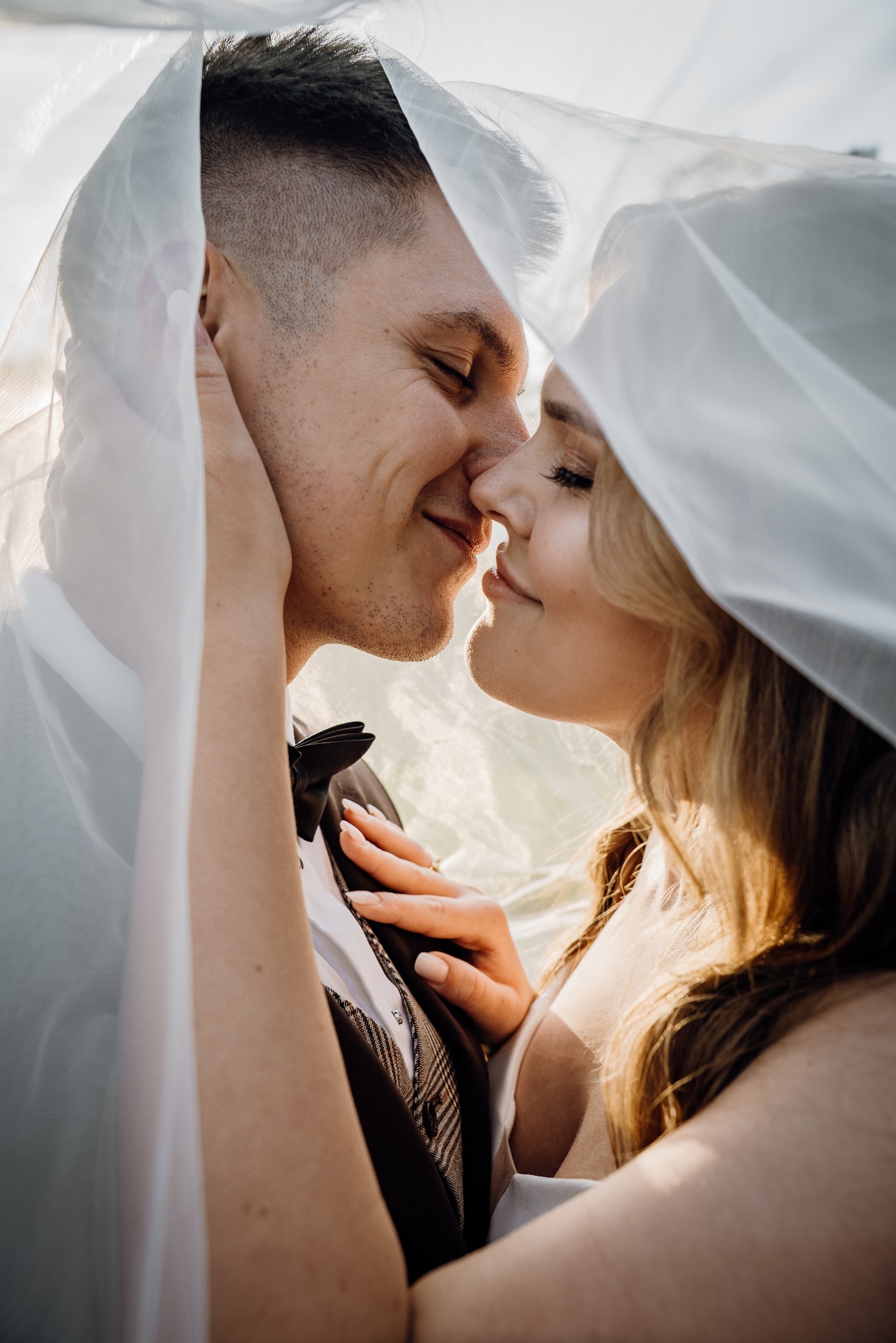 Młoda para w zbliżeniu, panna młoda w welonie, delikatne światło podkreśla rysy twarzy, subtelny uśmiech, elegancki strój pana młodego z muchą.